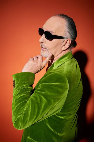 Pensive senior man with grey hair and beard, in stylish casual attire touching face and looking away on red and orange background with shadow, dark sunglasses, green velour blazer, fashion and age — Stock Photo
