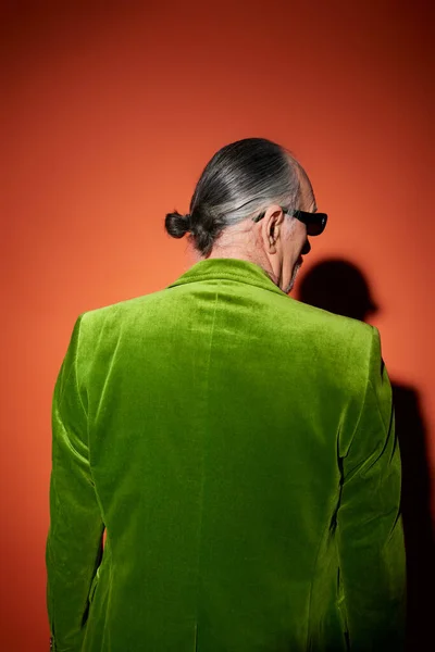 Back view of grey haired senior man in trendy casual clothes, green velour blazer and dark sunglasses, standing on red and orange background with shadow, fashionable senior male model — Stock Photo