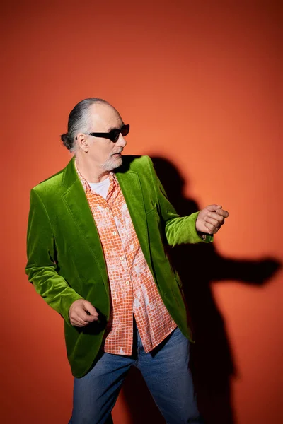 Cool homme âgé dans des lunettes de soleil sombres, veston en velours vert et chemise à la mode s'amuser, danser et regarder loin sur fond rouge et orange avec ombre, personnalité vibrante, concept de vieillissement heureux — Photo de stock