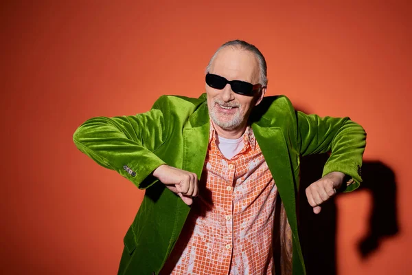 Joy and happiness, excited and fashionable senior man in dark sunglasses, trendy shirt and green velour blazer having fun and dancing on red and orange background with shadow — Stock Photo