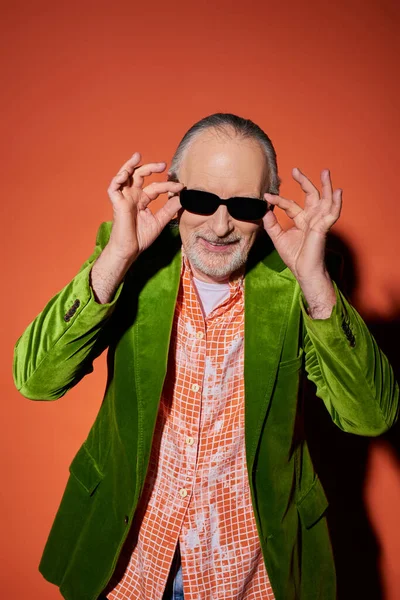 Optimistic senior man with groomed beard adjusting dark sunglasses and smiling at camera on red and orange background, fashion look, green velour blazer, happy and stylish aging concept — Stock Photo