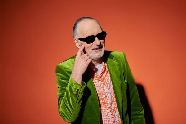 Homem idoso brincalhão e elegante apontando para a bochecha enquanto pede beijo no fundo vermelho e laranja, camisa na moda, blazer de veludo verde, óculos escuros, conceito de personalidade de envelhecimento vibrante — Fotografia de Stock