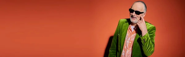 Cheerful senior man in dark sunglasses, trendy shirt and green velour blazer pointing at cheek and asking for kiss on red and orange background, happy aging, banner with copy space — Stock Photo