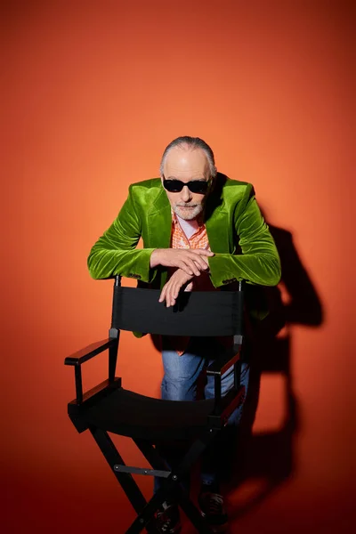 Vibrant and stylish senior personality, elderly man in dark sunglasses and green velour blazer leaning on chair and looking at camera on red and orange background with shadow, positive aging concept — Stock Photo