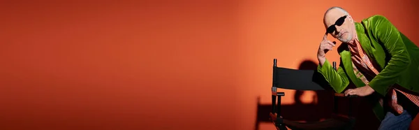 Nachdenkliches älteres männliches Modell in grünem Velours-Blazer und dunkler Sonnenbrille, das in die Kamera blickt, während es in der Nähe eines Stuhls auf rotem und orangefarbenem Hintergrund mit Schatten posiert, Modelook, Banner — Stockfoto