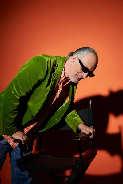 Heureux homme âgé en tenue élégante riant tout en se tenant près de la chaise, lunettes de soleil sombres, veston en velours vert, modèle plus ancien à la mode sur fond rouge et orange avec ombre — Photo de stock