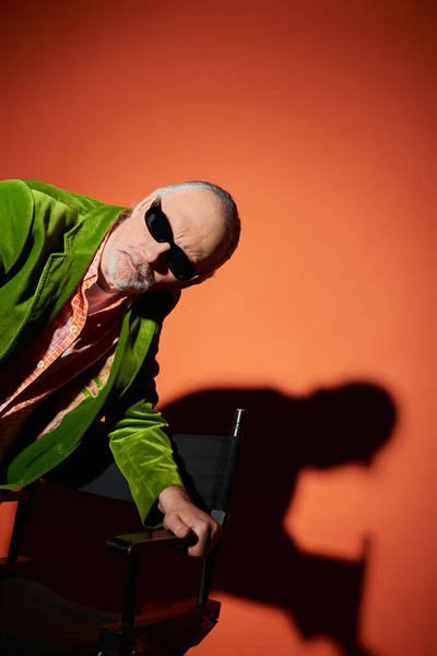 Hombre anciano reflexivo y curioso en gafas de sol oscuras y chaqueta de terciopelo verde de pie cerca de la silla y mirando hacia otro lado en el fondo rojo y naranja con sombra, envejecimiento de moda, estilo de vida elegante - foto de stock