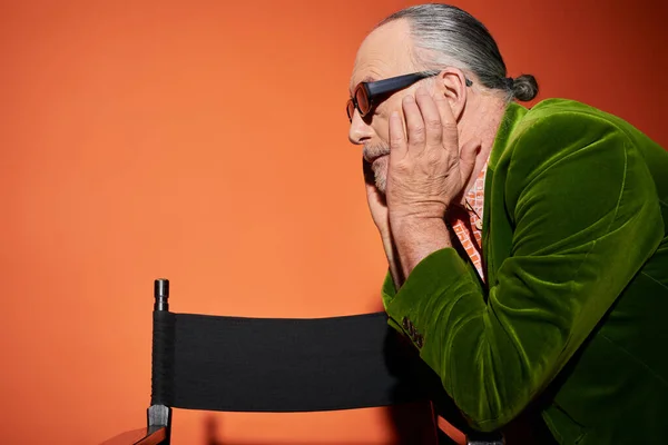 Side view of senior grey haired model touching face and looking away near chair on red and orange background, fashion look, dark sunglasses, green velour blazer, fashionable aging concept — Stock Photo