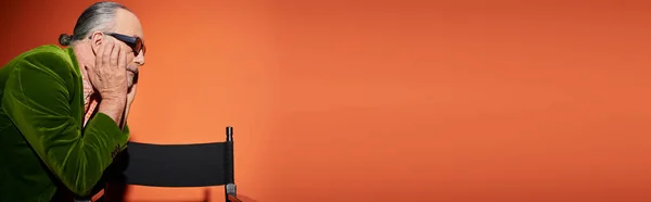 Вид сбоку старшей мужской модели в зеленом велюровом блейзере и темных солнцезащитных очках, стоящей рядом со стулом, трогательное лицо и размышления на красно-оранжевом фоне, личный стиль, повседневная мода, баннер — стоковое фото
