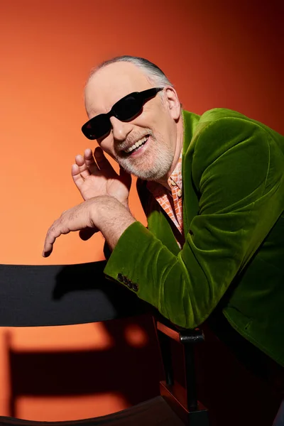 Hombre barbudo despreocupado y emocionado en traje elegante sonriendo cerca de la silla sobre fondo rojo y naranja, gafas de sol oscuras, chaqueta de terciopelo verde, concepto de envejecimiento feliz y de moda - foto de stock