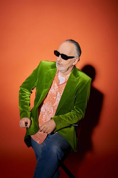 Älterer und selbstbewusster bärtiger Mann auf Stuhl sitzend und auf rotem und orangefarbenem Hintergrund mit Schatten wegschauend, Modelook, dunkle Sonnenbrille, trendiges Hemd, grüner Velours-Blazer, Mode- und Alterskonzept — Stockfoto
