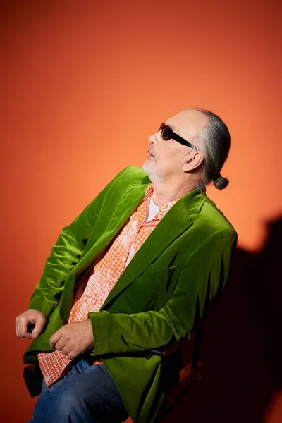 Hombre mayor sorprendido en gafas de sol oscuras, camisa de moda y chaqueta de terciopelo verde sentado en la silla y mirando hacia otro lado en el fondo rojo y naranja con sombra, look de moda, concepto de envejecimiento positivo - foto de stock