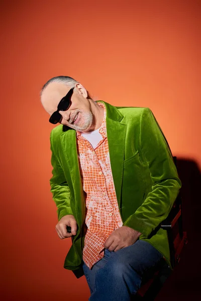 Joyful senior man with grey hair and beard sitting on chair and smiling at camera on red and orange background, dark sunglasses, green velour blazer, fashion trend, happy aging concept — Stock Photo