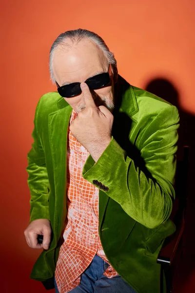 Ausdrucksstarker älterer Herr mit dunkler Sonnenbrille und Blick in die Kamera auf rotem und orangefarbenem Hintergrund mit Schatten, modischem Look, grünem Velours-Blazer, positiver und modischer Alterung — Stockfoto