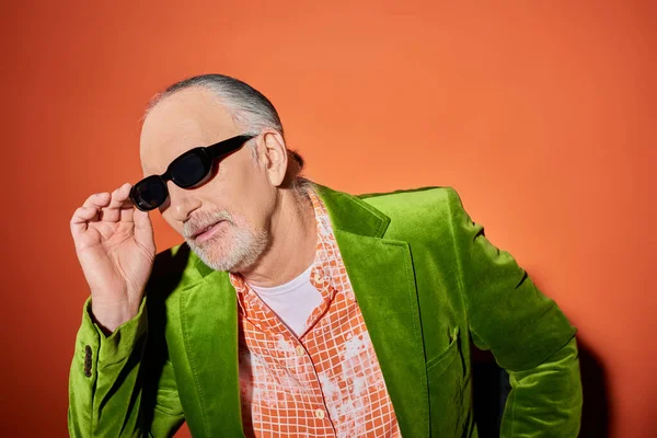 Individualidad vibrante, concepto de moda y edad, hombre barbudo senior con camisa de moda y chaqueta de terciopelo verde ajustando las gafas de sol oscuras mientras está sentado y mirando hacia otro lado en el fondo rojo y naranja - foto de stock