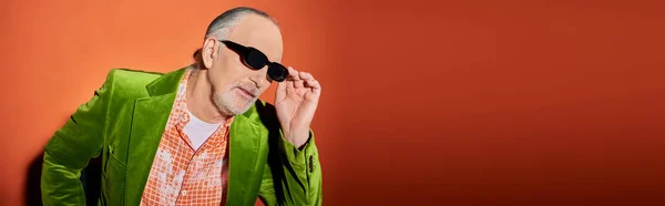 Senior man model in trendy shirt and green velour blazer touching dark sunglasses and looking away on red and orange background, stylish casual clothes, fashion and age concept, banner — Stock Photo