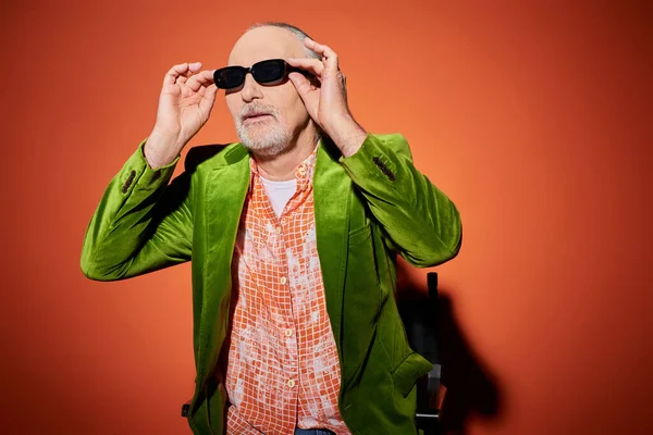Positive and fashionable aging concept, senior male model in green velour blazer and trendy shirt looking away and adjusting dark sunglasses while sitting on chair on red and orange background — Stock Photo