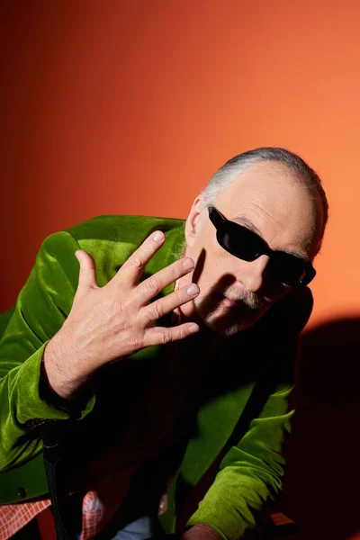 Expressive senior male model in dark sunglasses and green velour blazer gesturing with hand near face and looking at camera on red and orange background with shadow, vibrant aging personality — Stock Photo