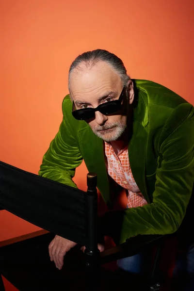Uomo anziano serio e fiducioso in giacca di velluto verde guardando la fotocamera sopra occhiali da sole scuri e alla moda mentre posa vicino alla sedia su sfondo rosso e arancione, concetto di invecchiamento alla moda — Foto stock