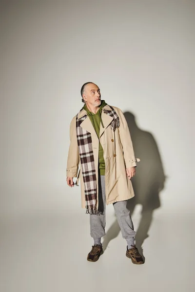 Full length of discouraged and thoughtful senior man in stylish casual clothes looking away on grey background with shadow, beige trench coat, green hoodie, plaid blazer, age positive concept — Stock Photo