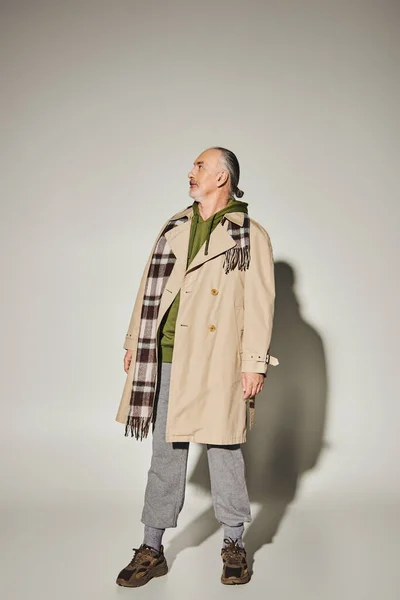 Longueur totale du modèle masculin senior en trench coat beige, écharpe à carreaux et sweat à capuche vert debout sur fond gris avec ombre et détournement des yeux, tenue décontractée élégante, concept de vieillissement à la mode — Photo de stock