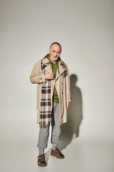 Fashion look, positive aging, full length of smiling senior male model in green hoodie, beige trench coat and plaid scarf standing and looking at camera on grey background with shadow — Stock Photo