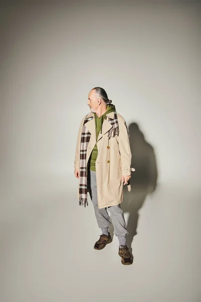 Повна довжина старого і бородатого чоловіка в бежевому стильному траншеї пальто, зелений светр і пухнастий шарф, що стоїть і дивиться на сірий фон з тіні і копіювати простір, моду і концепцію віку — стокове фото