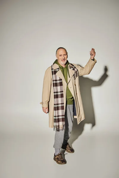 Full length of expressive and optimistic senior man in green hoodie, plaid scarf and beige trench coat gesturing and looking at camera on grey background with shadow, happy age concept — Stock Photo