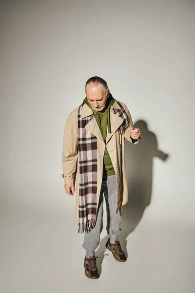 Full length of aged bearded man looking down while standing in trendy casual attire on grey background with shadow, beige trench coat, plaid scarf, green hoodie, fashion and age concept — Stock Photo
