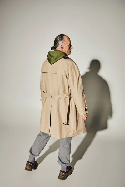 Вид ззаду на старшу модель в стильному повсякденному одязі, що стоїть на сірому фоні з тіні, у віці та сірому волоссі чоловік у бежевому траншеї пальто та зеленому светрі, концепція модного способу життя — стокове фото