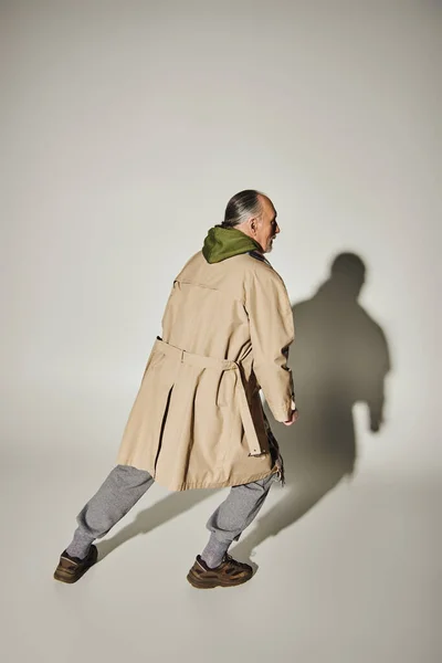 Langer älterer Mann in beigem Trenchcoat und grünem Kapuzenpullover, der in ausdrucksstarker Pose auf grauem Hintergrund mit Schatten steht, lässige Mode, stilvolles und positives Alterskonzept — Stockfoto