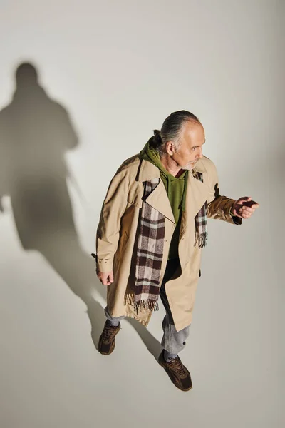 Повна довжина старшого чоловіка в стильному повсякденному вбранні, що стоїть на сірому фоні з тіні і дивиться вбік, зелений светр, бежеве траншеєве пальто, плетений шарф, модна концепція старіння, високий кут зору — стокове фото
