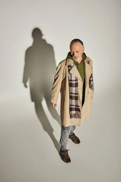 Vollbärtiger Mann in beigem Trenchcoat, grünem Kapuzenpullover und kariertem Schal, der auf grauem Hintergrund mit Schatten steht, trendige Freizeitkleidung, persönlicher Stil, Blickwinkel hoch — Stockfoto