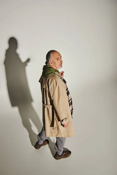 Homme âgé réfléchi en sweat à capuche vert, trench coat beige et blazer à carreaux regardant loin sur fond gris avec ombre, mode de vie à la mode, vue grand angle — Photo de stock