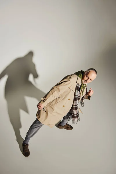 Top view of confident senior model in plaid scarf, green hoodie and beige trench coat standing in stylish pose on grey background with shadow, fashionable aging concept — Stock Photo
