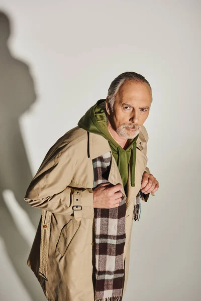 Confident aged man with serious and expressive gaze looking at camera on grey background with shadow, stylish casual wear, green hoodie, plaid scarf, beige trench coat, fashion and age concept — Stock Photo