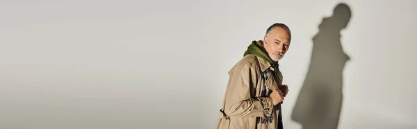 Concept de mode et d'âge, cher homme âgé en sweat à capuche vert, trench coat beige et écharpe à carreaux regardant loin tout en se tenant debout sur fond gris avec ombre, bannière avec espace de copie — Photo de stock