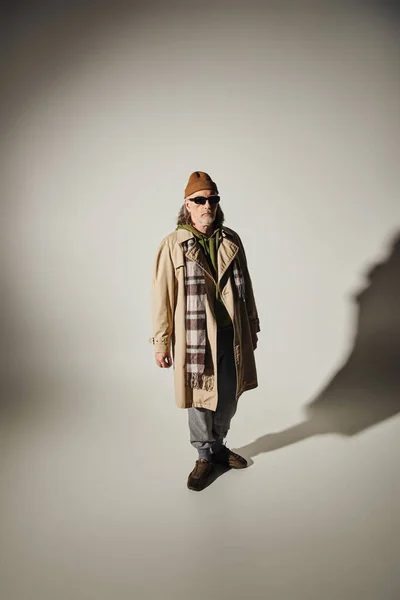 Mode- und Alterskonzept, hipsterhafter Senior mit Hut, dunkler Sonnenbrille, beigem Trenchcoat und kariertem Schal auf grauem Hintergrund mit Schatten und Kopierraum — Stockfoto