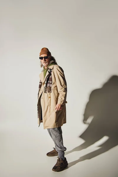 Полная длина модной старшей модели в темных солнцезащитных очках, шляпе-шапочке, бежевом плаще и клетчатом шарфе, стоящих на сером фоне с тенью, позитивной и модной концепцией старения — стоковое фото
