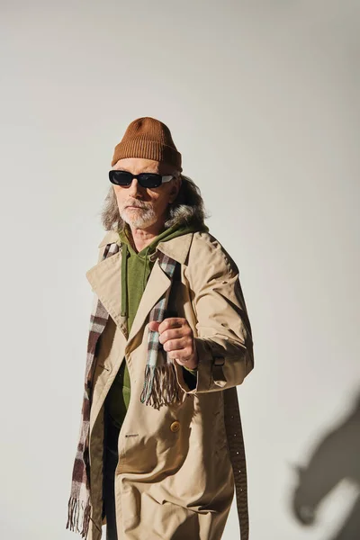 Confident senior man in dark sunglasses and hipster style attire standing with clenched fist and looking at camera on grey background, beanie hat, beige trench coat, fashionable aging concept — Stock Photo