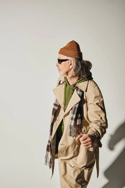 Стильна старша чоловіча модель, що стоїть і дивиться в сторону сірого фону, старий хіпстер в темних сонцезахисних окулярах, капелюх бані, бежеве тренч пальто і плетений шарф, концепція модного способу життя — стокове фото