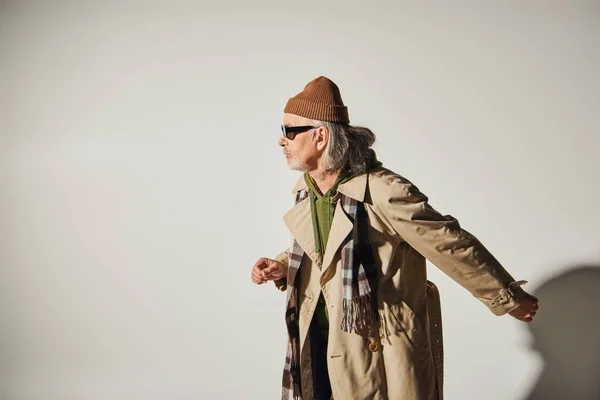 Vista lateral del hombre mayor en ropa casual de moda caminando sobre fondo gris, sombrero de gorro, gafas de sol oscuras, gabardina beige, bufanda a cuadros, estilo hipster, concepto de envejecimiento de moda - foto de stock