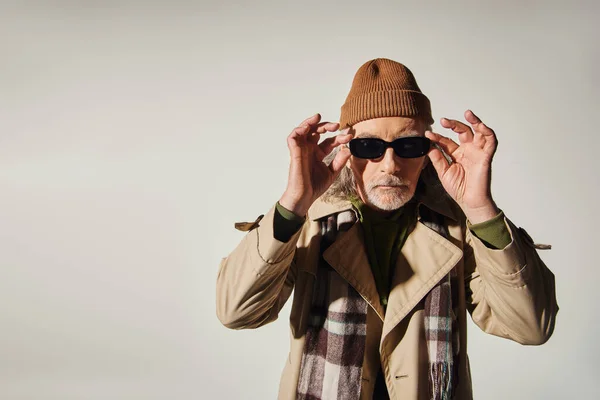 Modisches Altern, Hipster-Stil, cooler älterer Herr mit Mütze, kariertem Schal und Trenchcoat, dunkle Sonnenbrille und Blick in die Kamera auf grauem Hintergrund, Modeshooting — Stockfoto