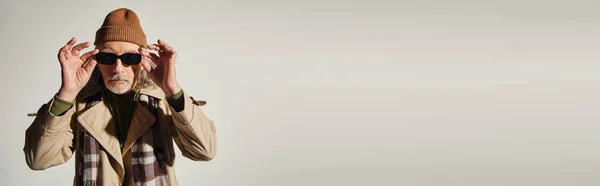 Homem sênior legal em chapéu de gorro, casaco de trincheira bege e cachecol xadrez ajustando óculos de sol na moda e olhando para a câmera em fundo cinza, moda hipster, envelhecimento elegante, banner com espaço de cópia — Fotografia de Stock