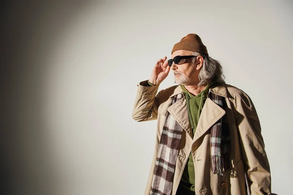 Anciano en gorro sombrero, gabardina beige y bufanda a cuadros ajustando gafas de sol oscuras y mirando hacia otro lado sobre fondo gris, estilo hipster, individualidad, moda y concepto de edad - foto de stock