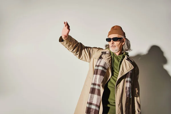 Modischer älterer Herr mit dunkler Sonnenbrille, Mütze, beigem Trenchcoat und kariertem Schal stehend mit ausgestrecktem und wegschauendem grauen Hintergrund, Hipster-Stil, ausdrucksstarke Persönlichkeit — Stockfoto
