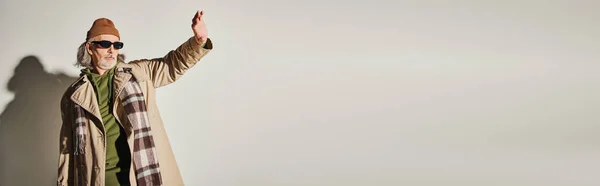 Homem de estilo hipster sênior na moda roupas casuais e óculos escuros de pé com a mão estendida e olhando para o fundo cinza, conceito de envelhecimento na moda, banner com espaço de cópia — Fotografia de Stock