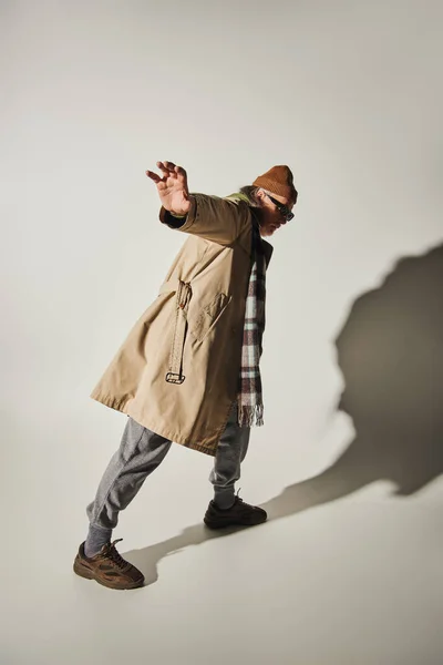 Full length of senior hipster man showing stop gesture while standing in expressive pose on grey background with shadow, dark sunglasses, beanie hat, beige trench coat, fashion and age concept — Stock Photo
