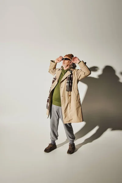 Full length of cool senior man in beanie hat and beige trench coat adjusting dark sunglasses and looking away on grey background with shadow, casual hipster style attire, fashion and age concept — Stock Photo