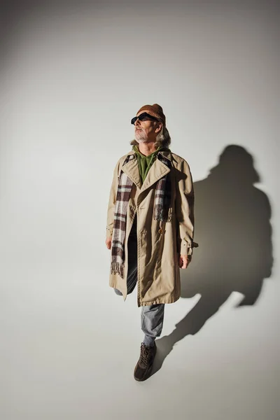 Hipster style, fashionable aging concept, high angle view of senior man in beanie hat, dark sunglasses, beige trench coat and plaid scarf posing on grey background with shadow — Stock Photo